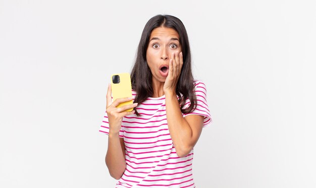 Young hispanic woman feeling shocked and scared and holding a smartphone