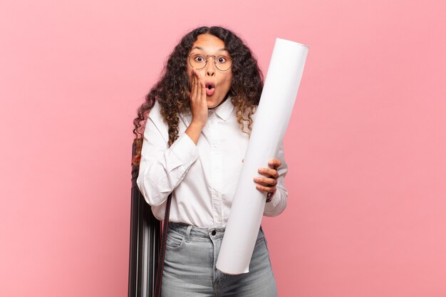 Young hispanic woman feeling shocked and scared, looking terrified with open mouth and hands on cheeks. architect concept