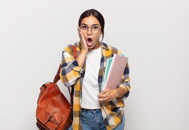 Young hispanic woman feeling shocked and scared, looking terrified with open mouth and hands on cheeks