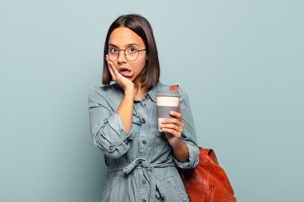 ショックを受けて怖がっている若いヒスパニック系の女性は、口を開けて頬に手を当てて恐怖を感じています