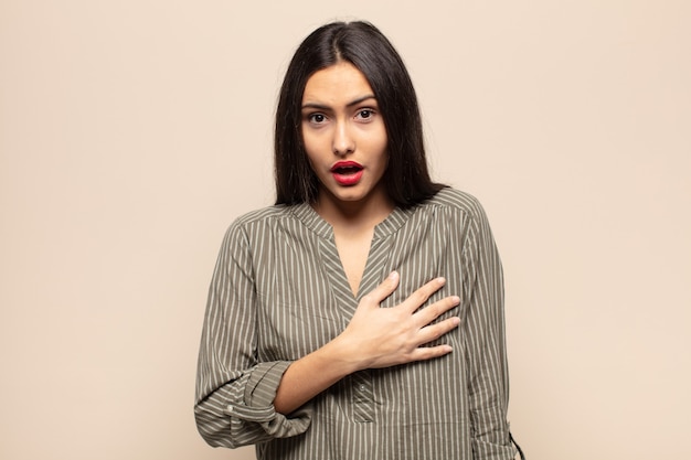 Young hispanic woman feeling shocked and surprised, smiling, taking hand to heart, happy to be the one or showing gratitude