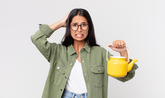Young hispanic woman feeling stressed anxious or scared with hands on head and holding a tea pot