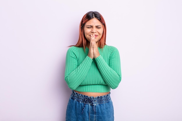 Young hispanic woman feeling worried, hopeful and religious, praying faithfully with palms pressed, begging forgiveness