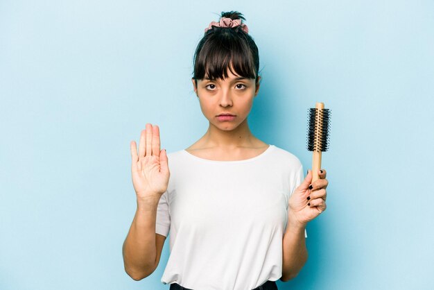 写真 青い背景に分離されたブラシの髪を保持している若いヒスパニック系の女性は、あなたを妨げる一時停止の標識を示す伸ばした手で立っています