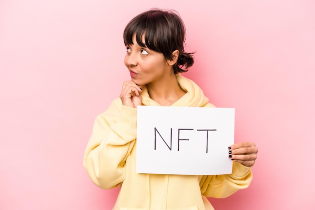 사진 분홍색 배경에 격리된 nft 플래카드를 들고 의심스럽고 회의적인 표정으로 옆을 바라보는 젊은 히스패닉 여성