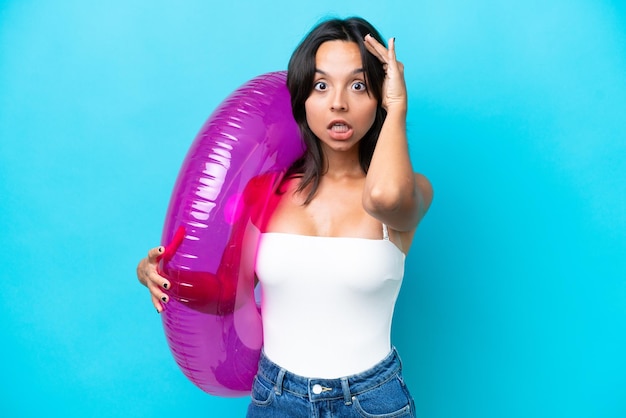 Young hispanic woman holding air mattress donut isolated on blue background doing surprise gesture while looking to the side