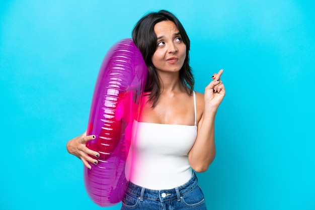 Young hispanic woman holding air mattress donut isolated on blue background with fingers crossing and wishing the best