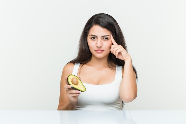 Young hispanic woman holding an avocado pointing his temple with finger, thinking, focused on a task