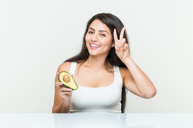 Giovane donna ispanica che tiene un avocado che mostra il segno di vittoria e che sorride ampiamente.