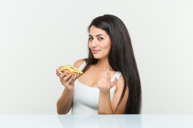 Young hispanic woman holding an avocado toast pointing with finger at you as if inviting come closer.