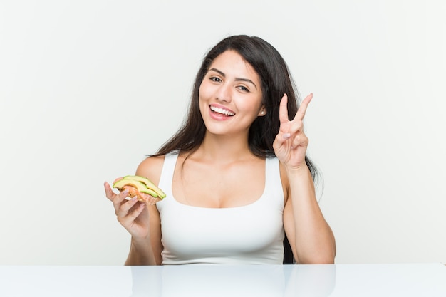 Young hispanic woman holding an avocado toast showing victory sign and smiling broadly.