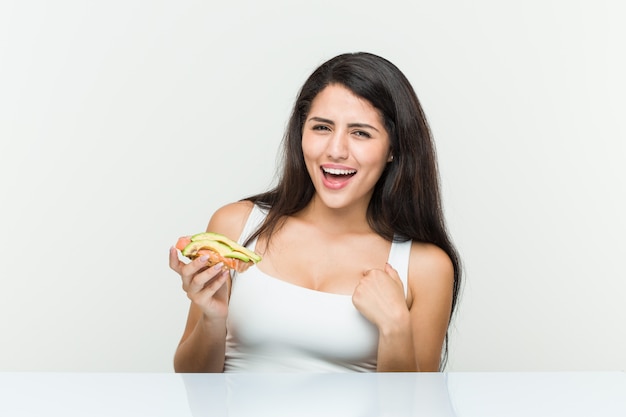 Young hispanic woman holding an avocado toast surprised pointing at himself, smiling broadly.