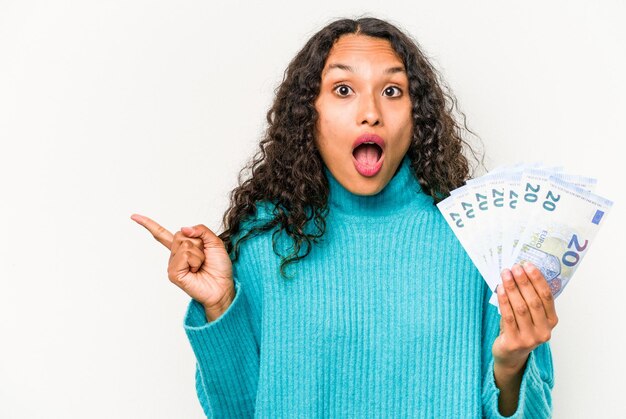 Young hispanic woman holding banknotes isolated on white background pointing to the side