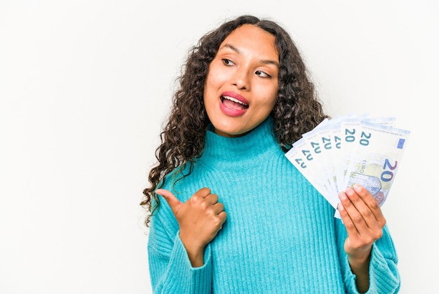 Young hispanic woman holding banknotes isolated on white background points with thumb finger away laughing and carefree