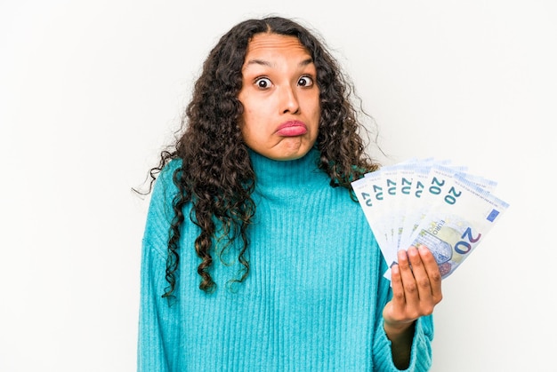 흰색 배경에 격리된 지폐를 들고 있는 젊은 히스패닉계 여성은 어깨를 으쓱하고 혼란스러운 눈을 뜨고 있다