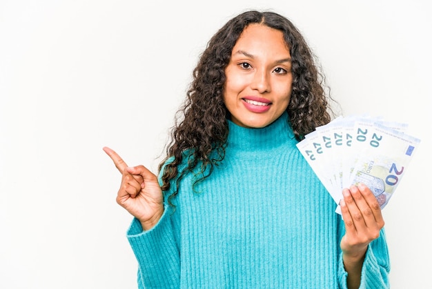 Young hispanic woman holding banknotes isolated on white background smiling and pointing aside showing something at blank space