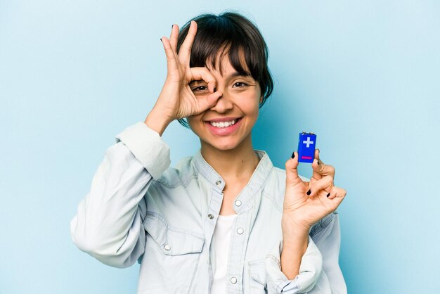 Young hispanic woman holding a batterie isolated on blue background excited keeping ok gesture on eye