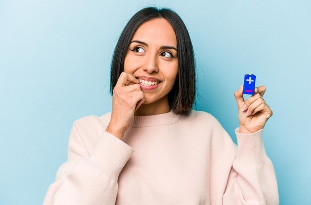 青い背景に分離された電池を保持している若いヒスパニック系の女性は、コピースペースを見ている何かについて考えてリラックスしました