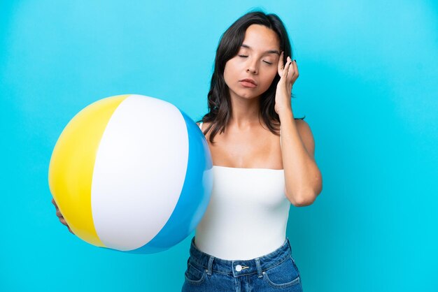 Young hispanic woman holding beach ball isolated on blue background with headache