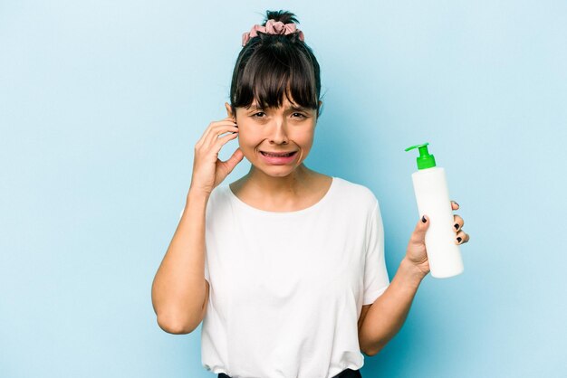 Young hispanic woman holding a body lotion isolated on blue background covering ears with hands