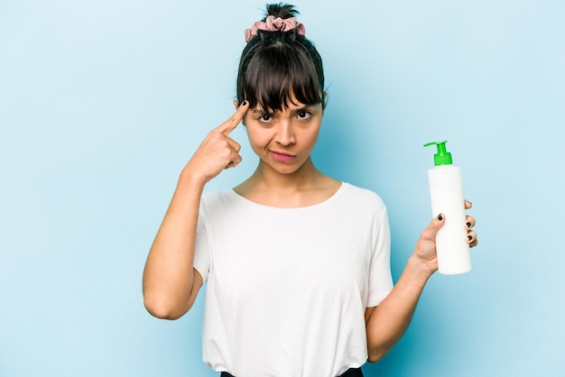 Young hispanic woman holding a body lotion isolated on blue background pointing temple with finger thinking focused on a task