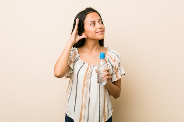 Foto giovane donna ispanica che tiene una bottiglia di acqua cercando di ascoltare un gossip.