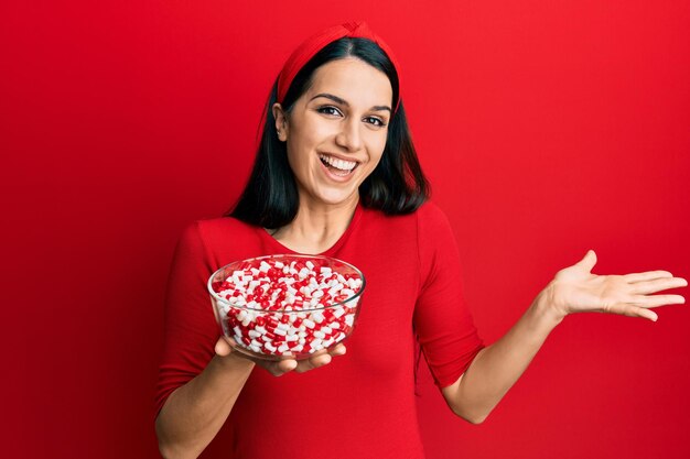 Young hispanic woman holding bowl of pills celebrating achievement with happy smile and winner expression with raised hand