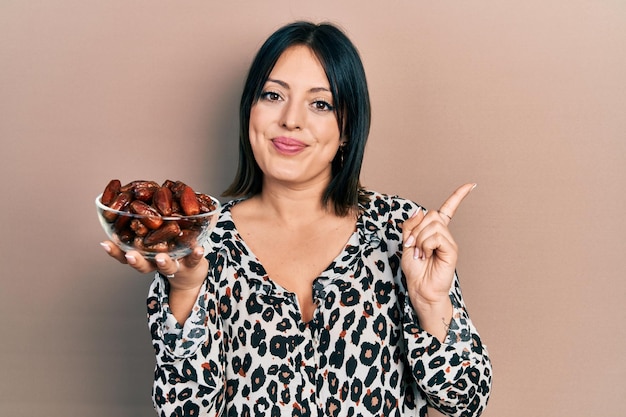 Young hispanic woman holding bowl with dates smiling happy pointing with hand and finger to the side