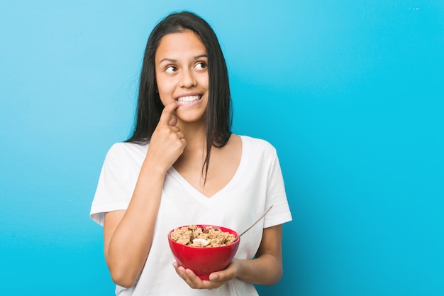 Young hispanic woman holding a cereal bowl relaxed thinking about something looking at a copy space.