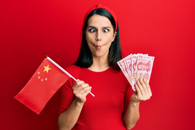 Young hispanic woman holding china flag and yuan banknotes making fish face with mouth and squinting eyes crazy and comical