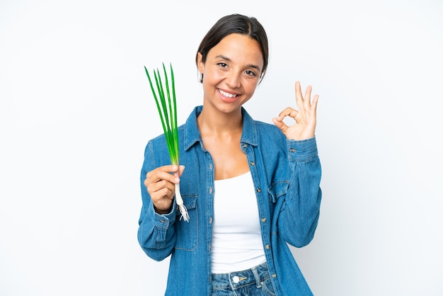 Young hispanic woman holding chive isolated on white background showing ok sign with fingers