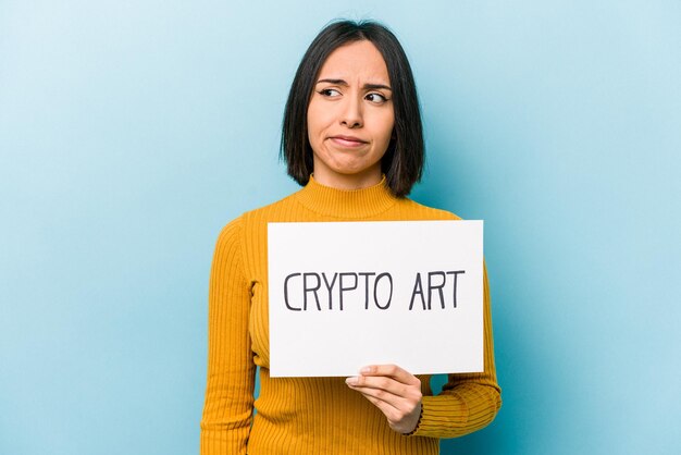 Young hispanic woman holding crypto art placard isolated on blue background confused feels doubtful and unsure