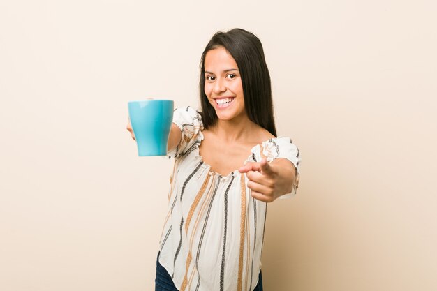 Young hispanic woman holding a cup cheerful smiles pointing to front.