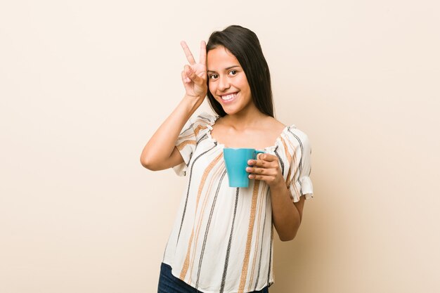 Young hispanic woman holding a cup showing victory sign and smiling broadly