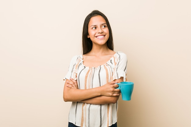 Young hispanic woman holding a cup smiling confident with crossed arms.