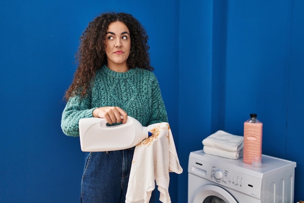 Young hispanic woman holding dirty laundry and detergent bottle smiling looking to the side and staring away thinking