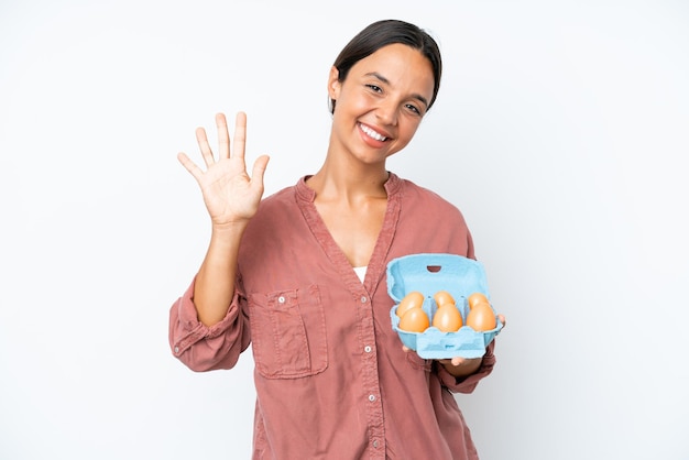 Молодая латиноамериканка держит яйца на белом фоне, считая пять пальцами