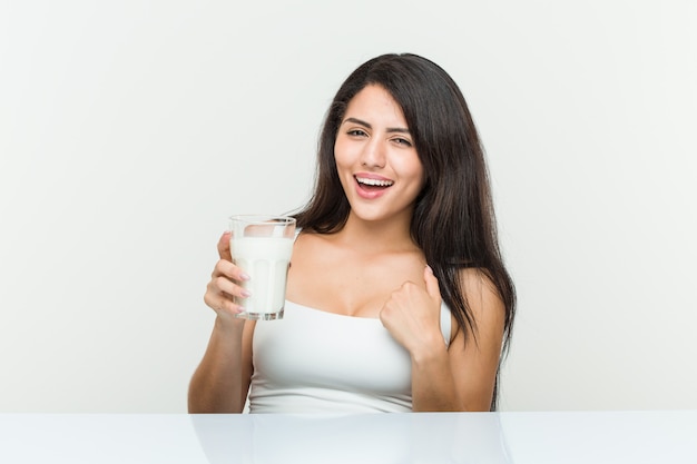 Young hispanic woman holding a glass of milk surprised pointing at herself