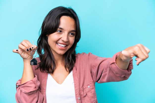 Photo young hispanic woman holding home keys isolated on blue background giving a thumbs up gesture