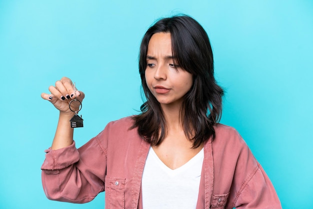 Photo young hispanic woman holding home keys isolated on blue background looking to the side