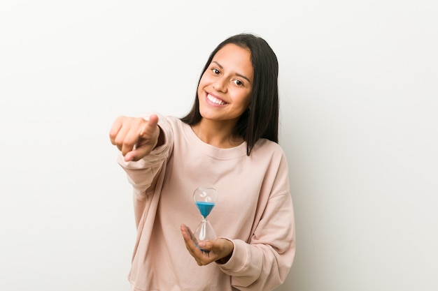 Young hispanic woman holding an hourglass cheerful smiles pointing to front.