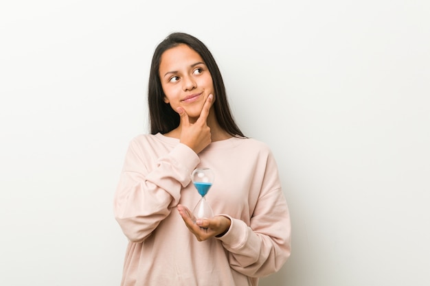 Young hispanic woman holding an hourglass looking sideways with doubtful and skeptical expression.