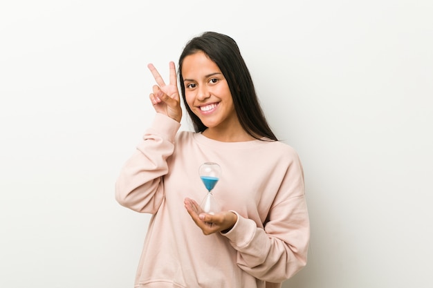 Young hispanic woman holding an hourglass showing victory sign and smiling broadly.