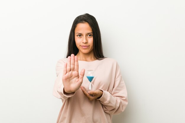 Young hispanic woman holding an hourglass standing with outstretched hand showing stop sign, preventing you.