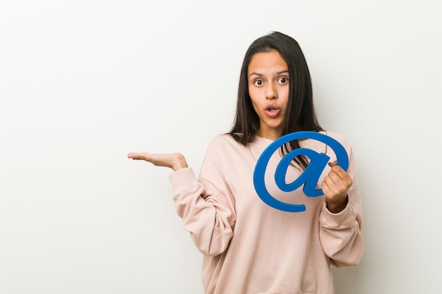 Young hispanic woman holding an at icon impressed holding copy space on palm.