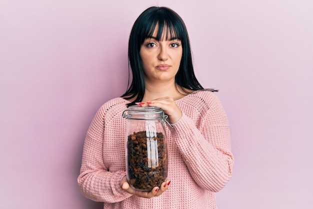 Young hispanic woman holding jar of raisins relaxed with serious expression on face. simple and natural looking at the camera.