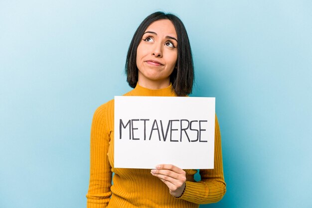 Young hispanic woman holding metaverse placard isolated on blue background dreaming of achieving goals and purposes