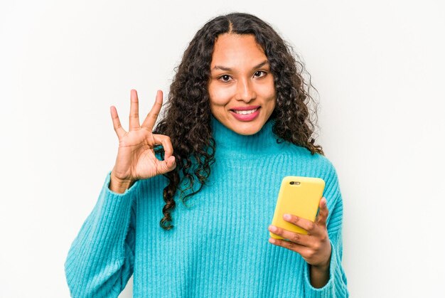 白い背景で隔離の携帯電話を保持している若いヒスパニック系女性陽気で自信を持ってOKジェスチャーを示しています