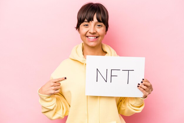 Young hispanic woman holding a NFT placard isolated on pink background smiling and pointing aside showing something at blank space