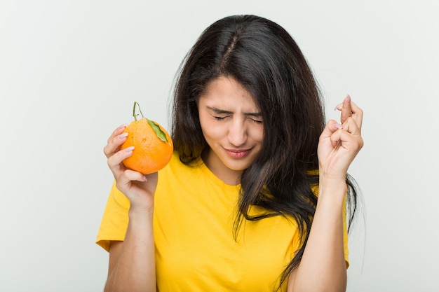 Young hispanic woman holding an orange crossing fingers for having luck